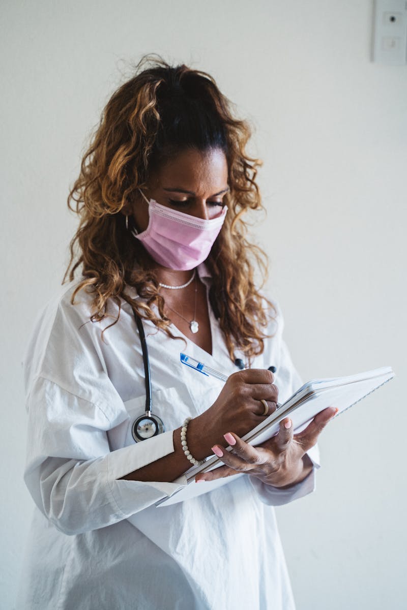 A female doctor wearing a mask writes notes in a clinical setting. Professional healthcare imagery.